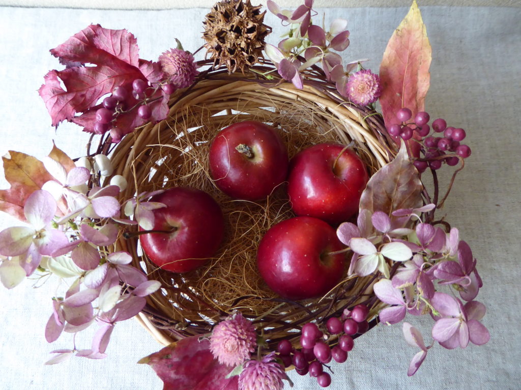 姫リンゴの入った丸いカゴのフチ回りに落ち葉や実物、ドライ素材を飾り付けたリース風アレンジの見本写真